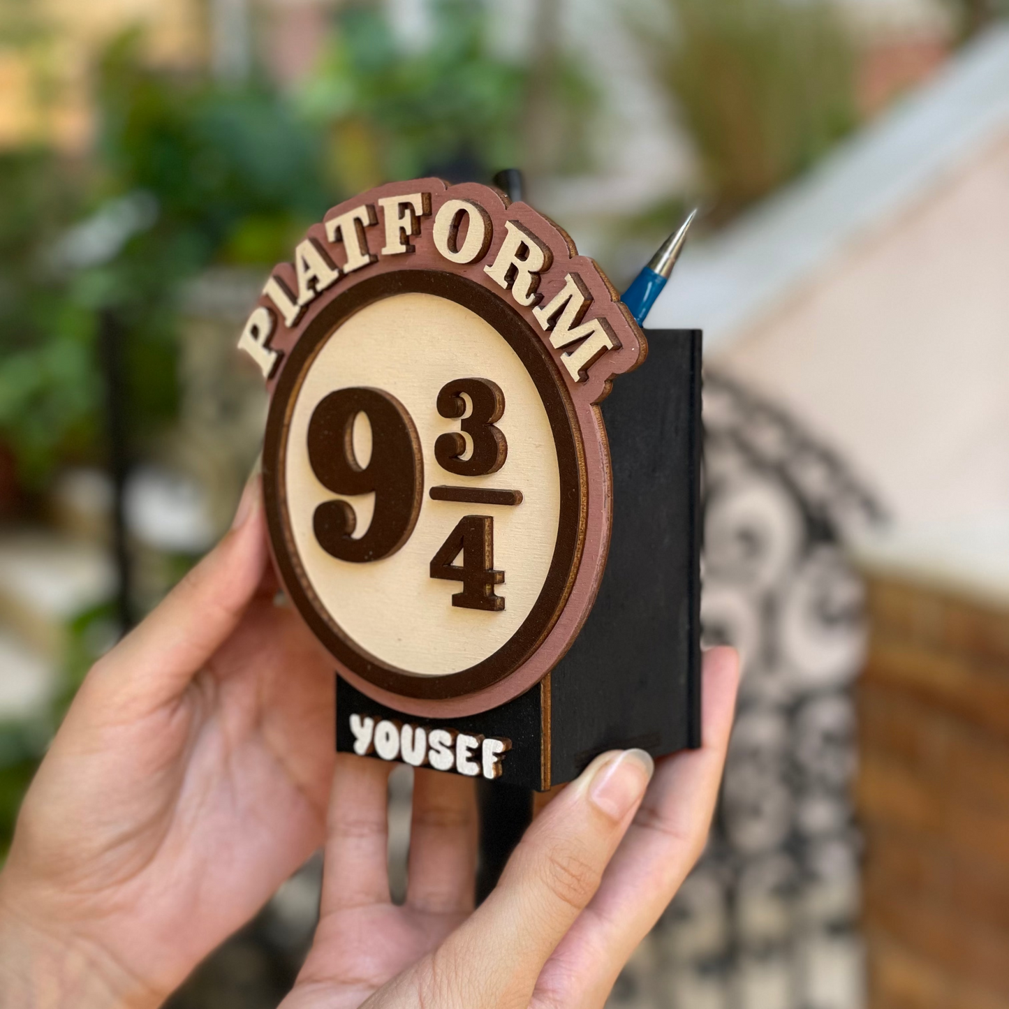 Hand holding a wooden bookmark with 'Platform 9 3/4' design and 'Yousef' engraving.