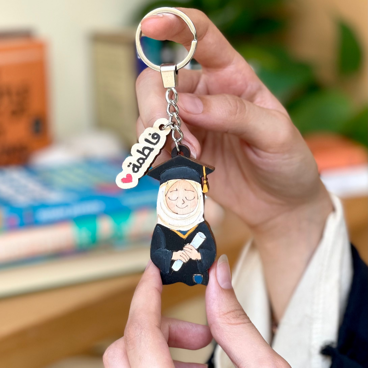 Person holding a keychain with a character and 'addio' charm in front of books and plants.