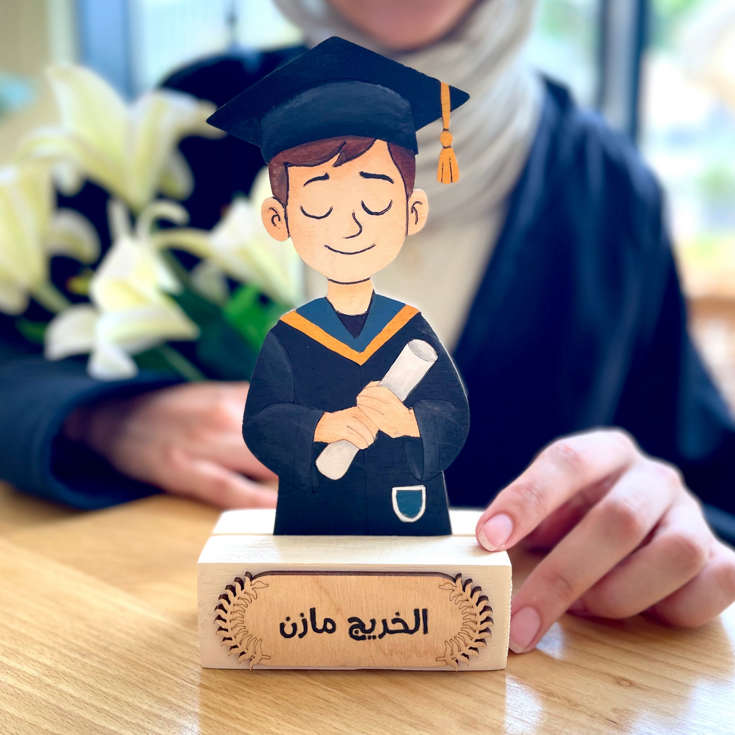 Graduation-themed figurine on a wooden stand with a person in the background.