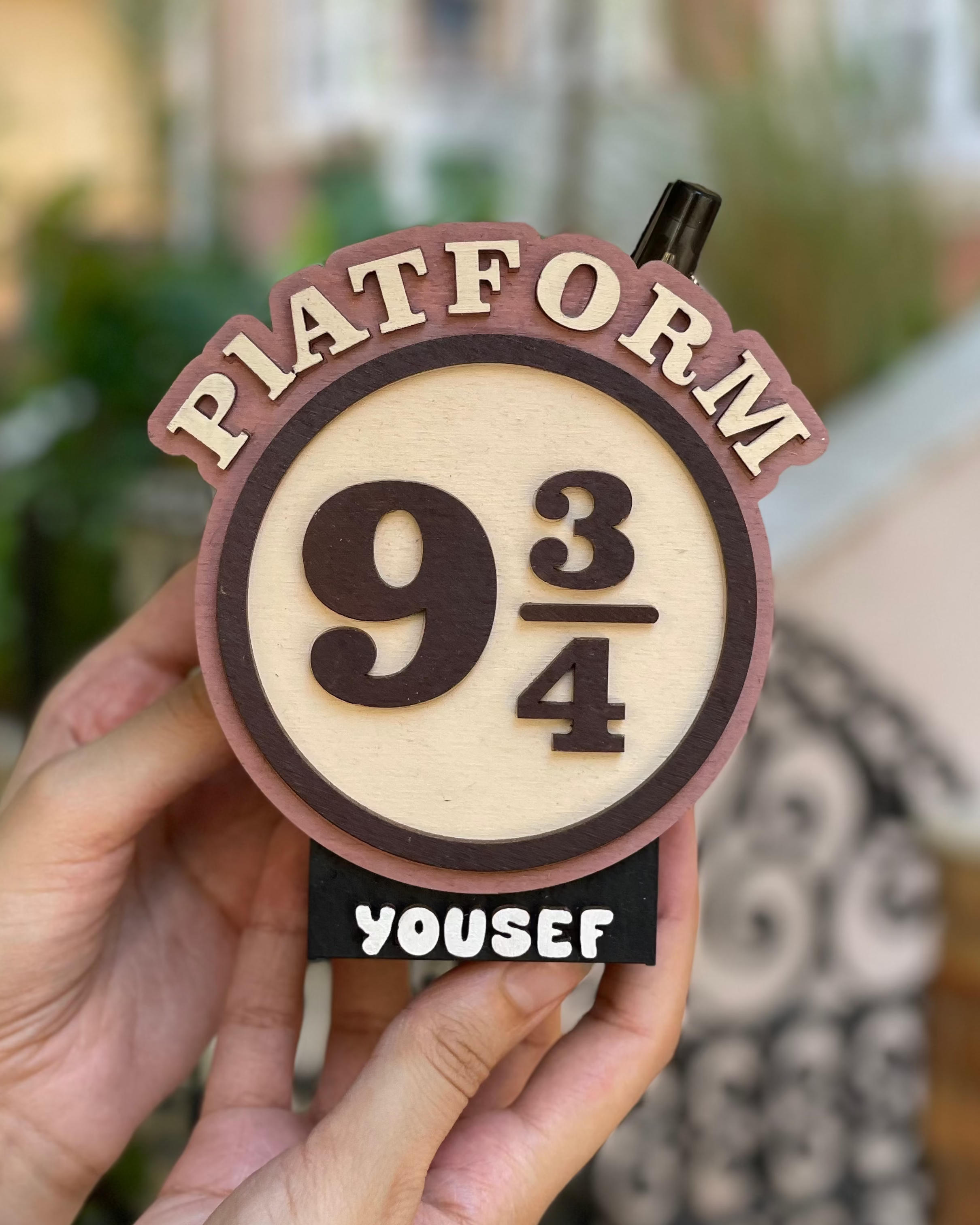 Hand holding a 'Platform 9 3/4' sign with 'Yousef' personalized name, blurred outdoor background
