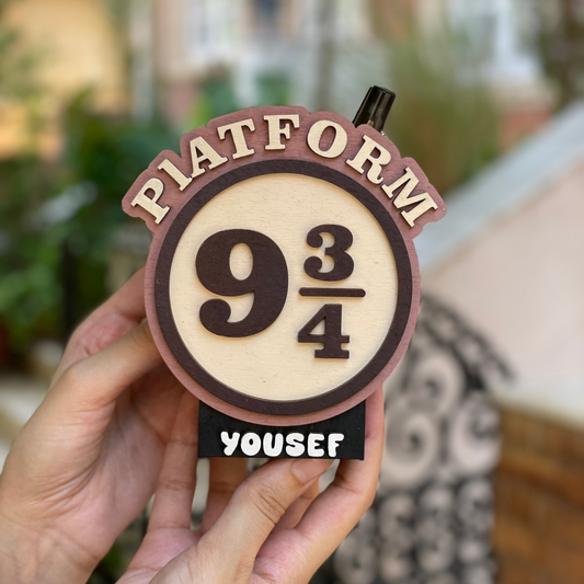 Hand holding a 'Platform 9 3/4' sign with 'Yousef' personalized name, blurred outdoor background