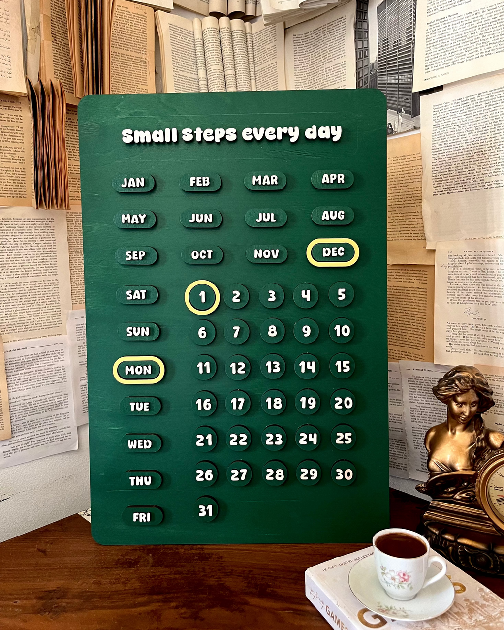 Green calendar with 'Small steps every day' text on a wooden surface with books and a cup of coffee in the background.