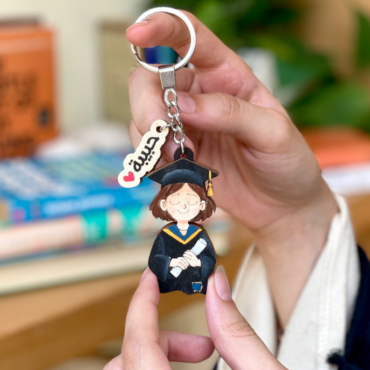 Graduation-themed keychain held by a person with a blurred indoor background