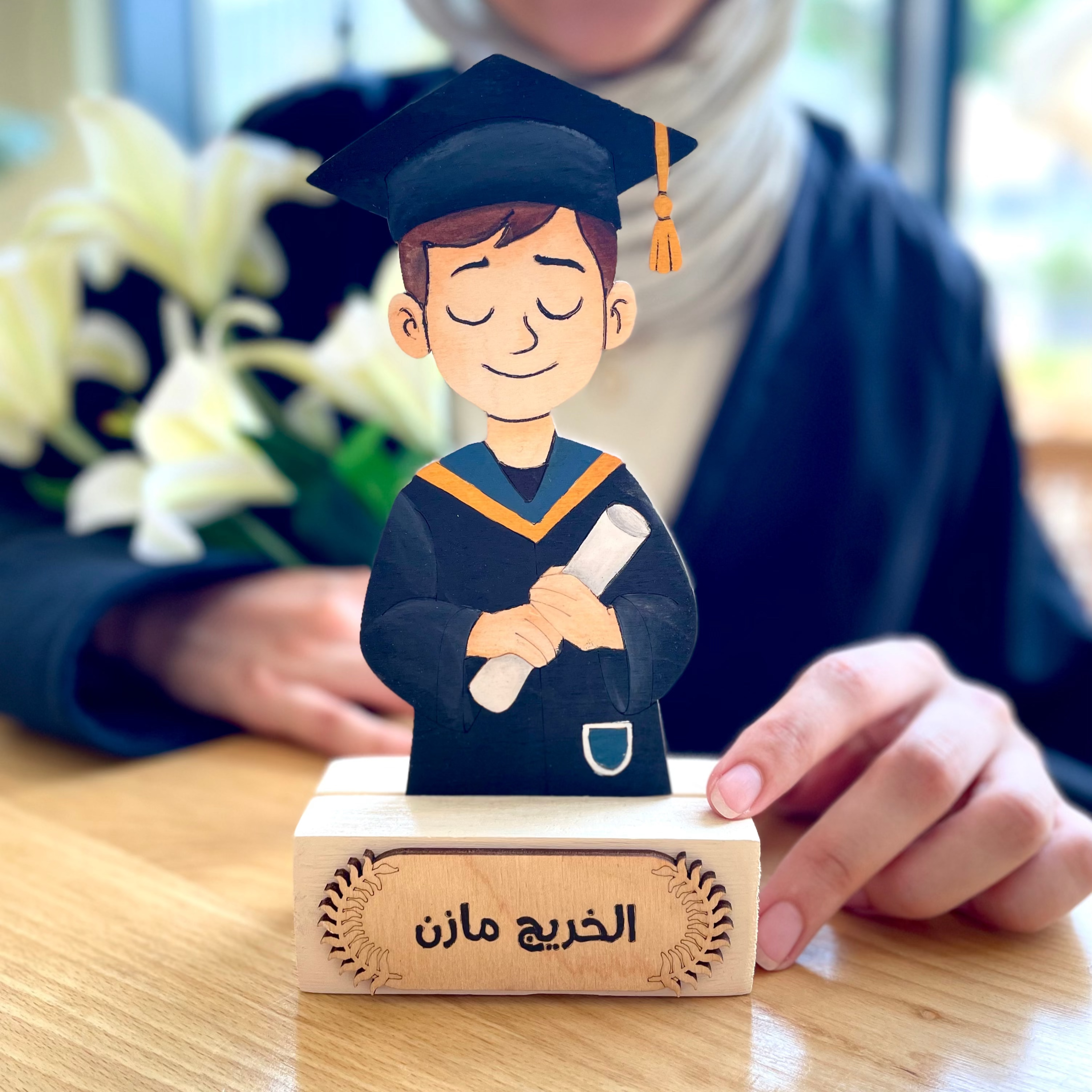 Graduation-themed figurine on a wooden stand with a person in the background.
