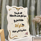 An off-white decorative plaque with Arabic graduation-themed text, gold script accents, and small black speckles, displayed on a table in front of sheer curtains with flowers nearby.