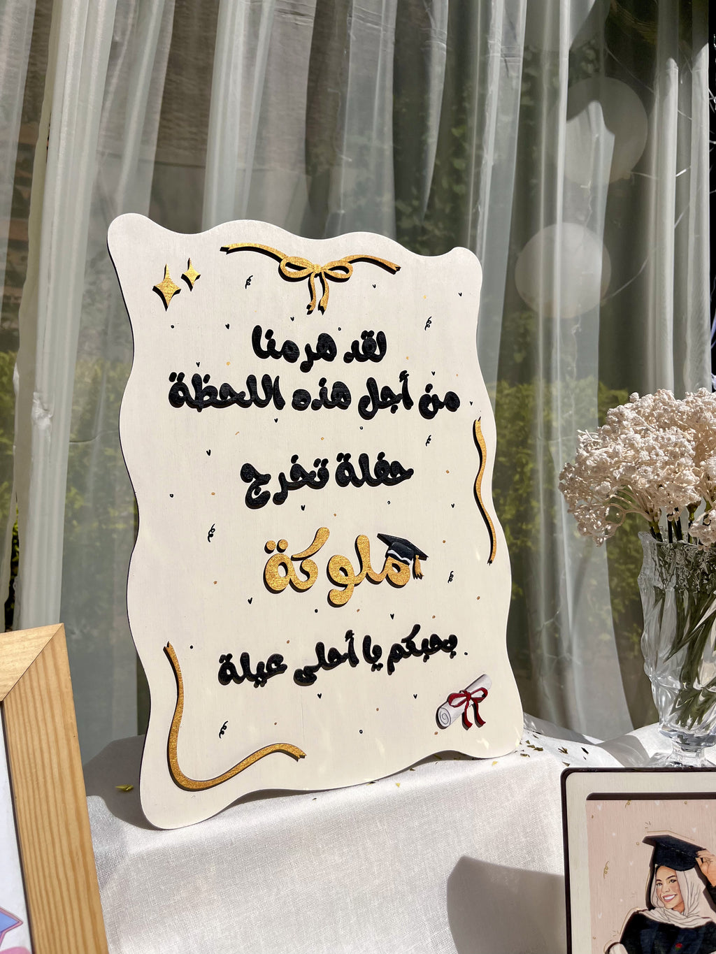 An off-white decorative plaque with Arabic graduation-themed text, gold script accents, and small black speckles, displayed on a table in front of sheer curtains with flowers nearby.