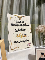 An off-white decorative plaque with Arabic graduation-themed text, gold script accents, and small black speckles, displayed on a table in front of sheer curtains with flowers nearby.