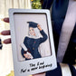 A person in a graduation gown holds a custom white square frame featuring an illustration of a woman in a graduation cap and gown with the text 'The End For a new beginning'.