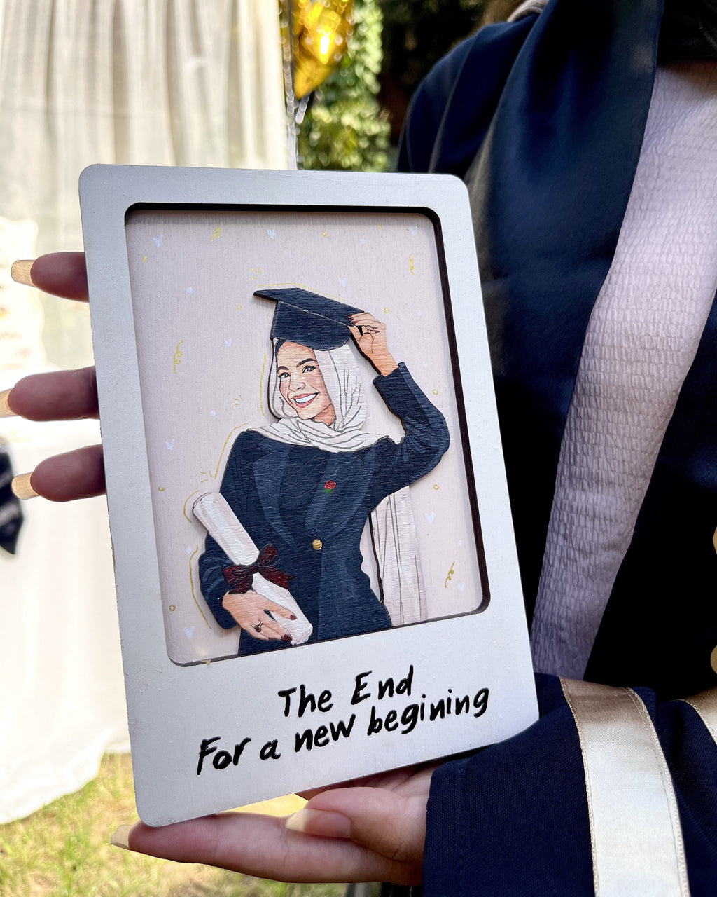 A person in a graduation gown holds a custom white square frame featuring an illustration of a woman in a graduation cap and gown with the text 'The End For a new beginning'.