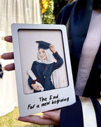 A person in a graduation gown holds a custom white square frame featuring an illustration of a woman in a graduation cap and gown with the text 'The End For a new beginning'.