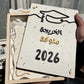 A hand holds a square wooden graduation gift box containing a decorative plaque with Arabic text reading 'تمكّك التخرج 2026' and a graduation cap illustration. The box is partially open, showing white ribbons inside, with other graduation-themed items blurred in the background.