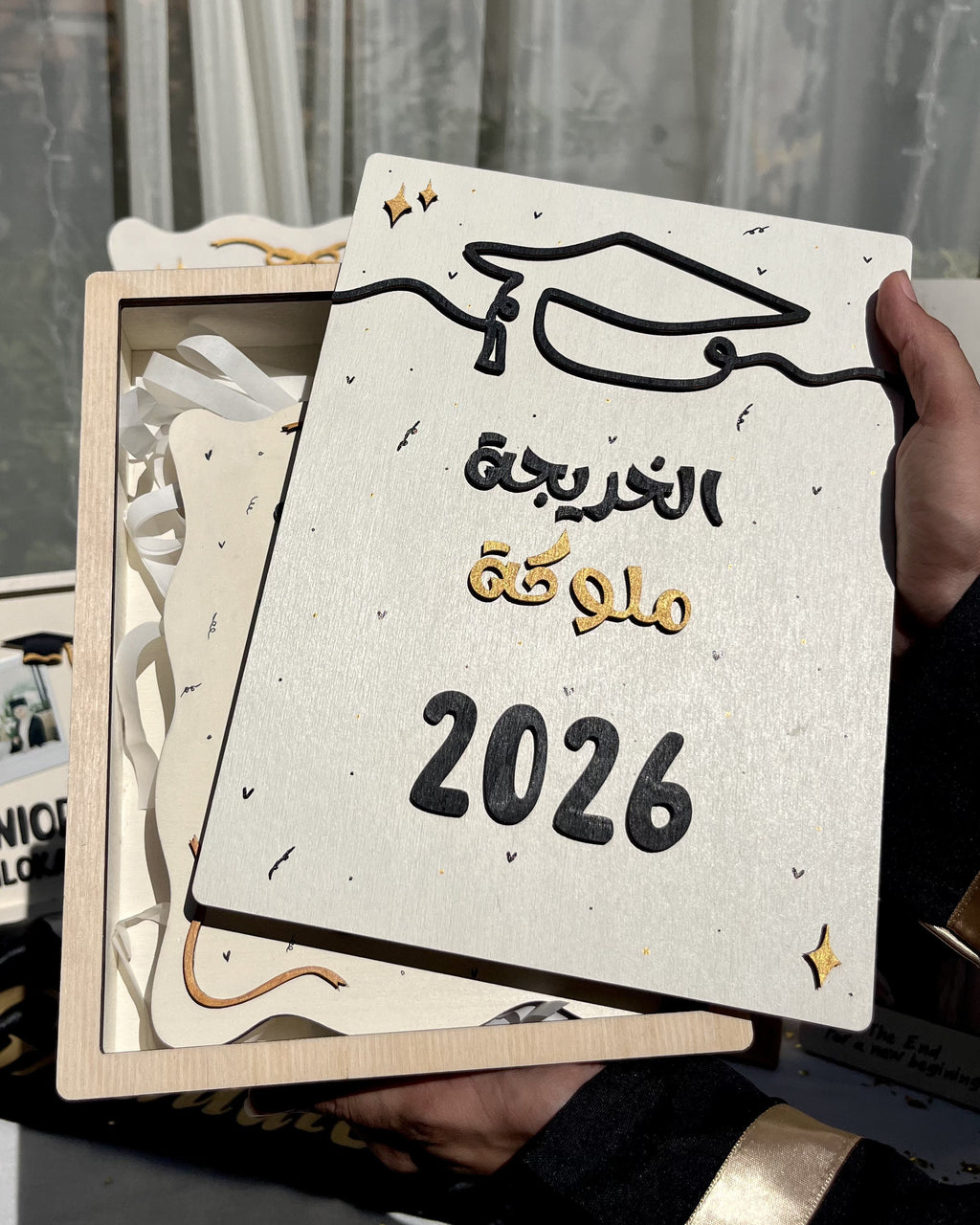 A hand holds a square wooden graduation gift box containing a decorative plaque with Arabic text reading 'تمكّك التخرج 2026' and a graduation cap illustration. The box is partially open, showing white ribbons inside, with other graduation-themed items blurred in the background.