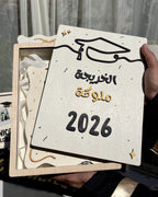 A hand holds a square wooden graduation gift box containing a decorative plaque with Arabic text reading 'تمكّك التخرج 2026' and a graduation cap illustration. The box is partially open, showing white ribbons inside, with other graduation-themed items blurred in the background.