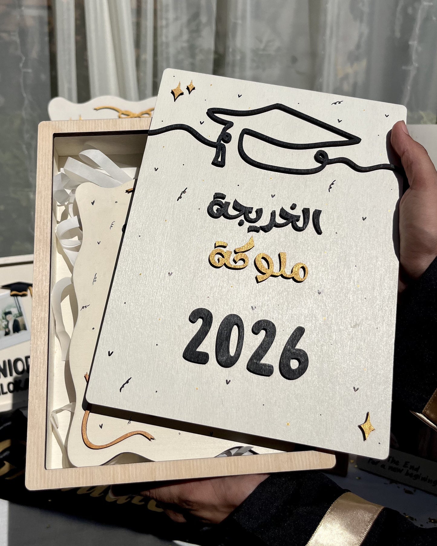 A hand holds a square wooden graduation gift box containing a decorative plaque with Arabic text reading 'تمكّك التخرج 2026' and a graduation cap illustration. The box is partially open, showing white ribbons inside, with other graduation-themed items blurred in the background.