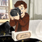 A custom wooden photo plaque featuring a cartoon illustration of a man holding a camera, mounted on a white rectangular base with Arabic text. The plaque is styled on a black satin fabric with gold confetti, with a photo collage and a white curtain in the background.