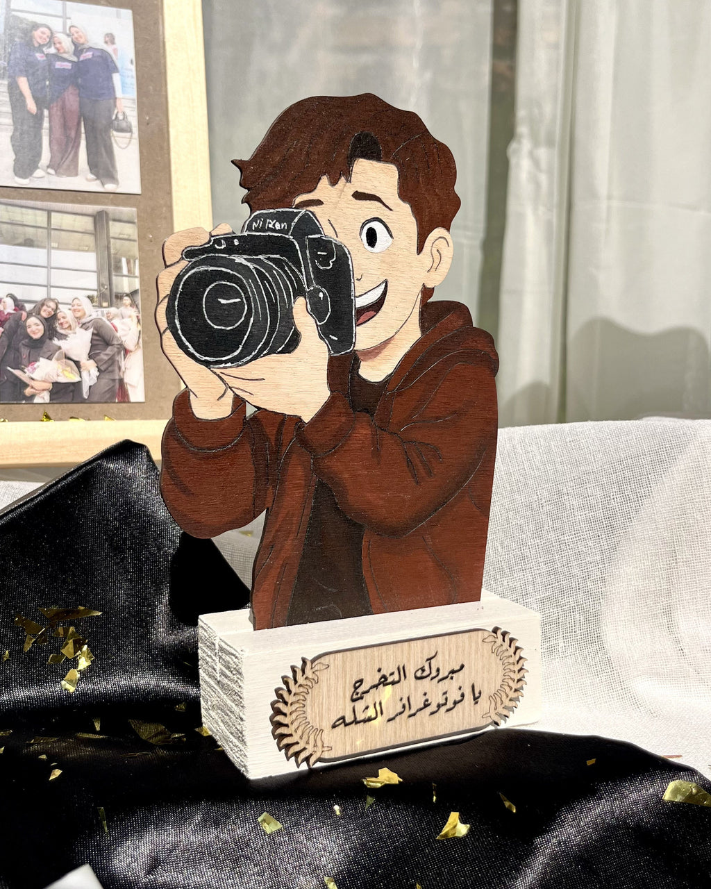A custom wooden photo plaque featuring a cartoon illustration of a man holding a camera, mounted on a white rectangular base with Arabic text. The plaque is styled on a black satin fabric with gold confetti, with a photo collage and a white curtain in the background.