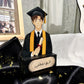 A personalized wooden graduation figurine featuring a cartoon-style graduate in a black gown and mortarboard cap with a gold sash, holding a diploma scroll. The base is black with Arabic calligraphy and a beige nameplate.