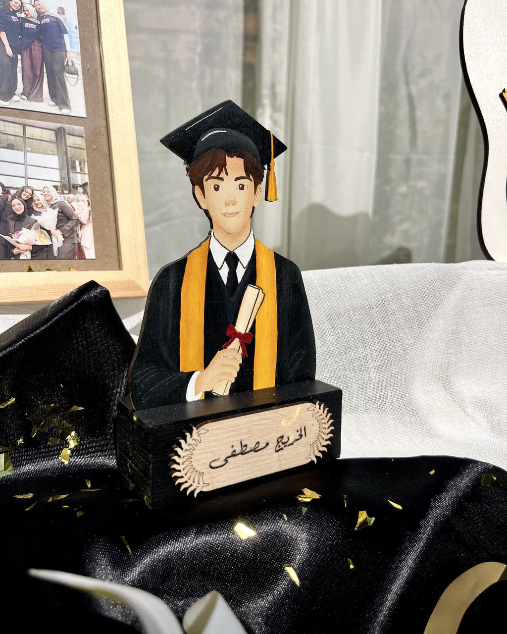 A personalized wooden graduation figurine featuring a cartoon-style graduate in a black gown and mortarboard cap with a gold sash, holding a diploma scroll. The base is black with Arabic calligraphy and a beige nameplate.