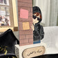 A personalized wooden keepsake box or plaque featuring an illustrated figure in a black hoodie and sunglasses peeking from behind a brick wall with sticky notes, set on a white base with Arabic text. The product is styled on black satin fabric with gold confetti, with a framed photo and a white curtain in the background.