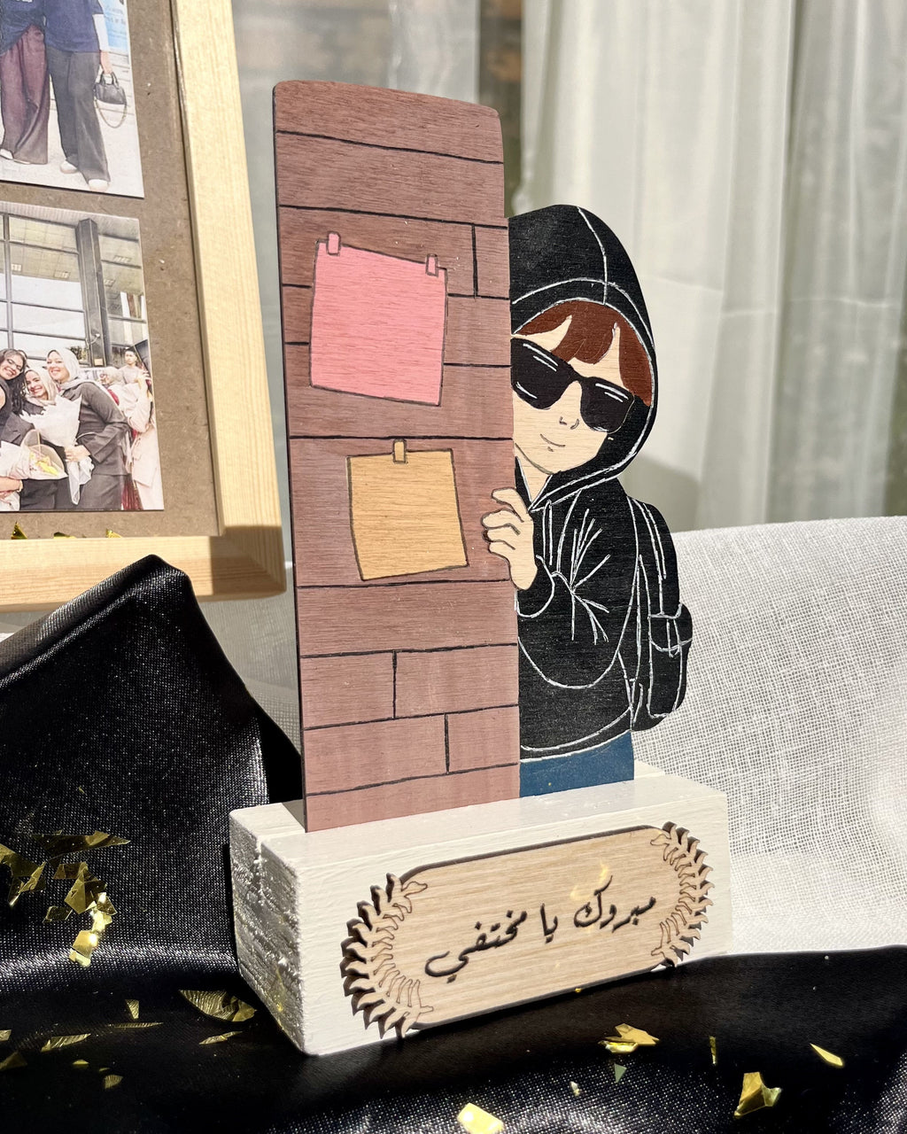 A personalized wooden keepsake box or plaque featuring an illustrated figure in a black hoodie and sunglasses peeking from behind a brick wall with sticky notes, set on a white base with Arabic text. The product is styled on black satin fabric with gold confetti, with a framed photo and a white curtain in the background.