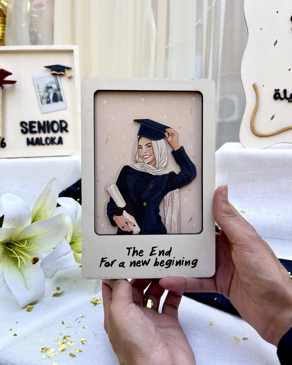 A person holds a square cream-colored keepsake photo frame featuring a 3D paper-cut illustration of a graduation scene with the text 'The End. For a new beginning'.