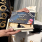 A person holds an open wooden book or presentation box containing a dark blue certificate with Arabic text, alongside a cream-colored diploma or award card. The scene appears to be outdoors near white curtains, with part of a black sash or garment visible on the left and a white banner with gold lettering below.
