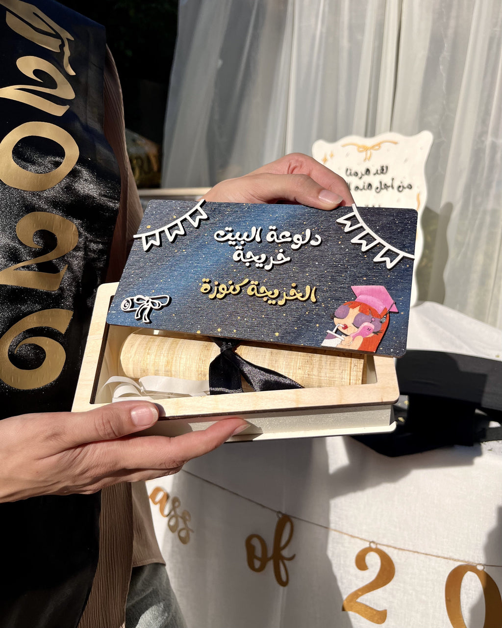 A person holds an open wooden book or presentation box containing a dark blue certificate with Arabic text, alongside a cream-colored diploma or award card. The scene appears to be outdoors near white curtains, with part of a black sash or garment visible on the left and a white banner with gold lettering below.