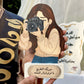A person holds a decorative wooden plaque featuring an illustrated woman holding a camera, with Arabic text on the plaque. Another similar plaque is visible in the background, along with a dark navy sash with gold lettering.