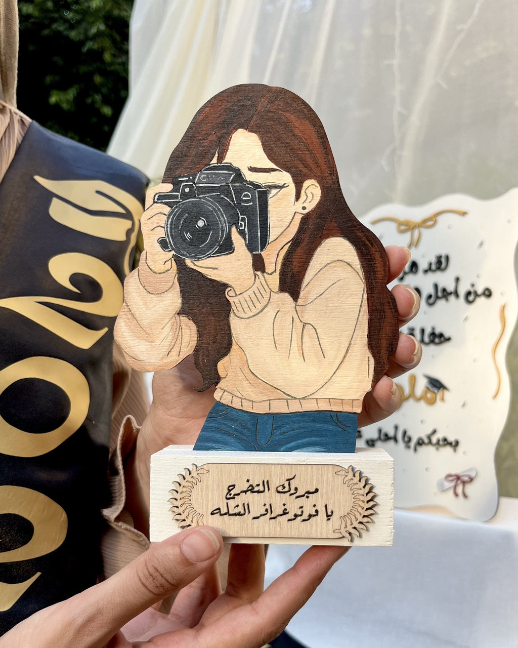 A person holds a decorative wooden plaque featuring an illustrated woman holding a camera, with Arabic text on the plaque. Another similar plaque is visible in the background, along with a dark navy sash with gold lettering.